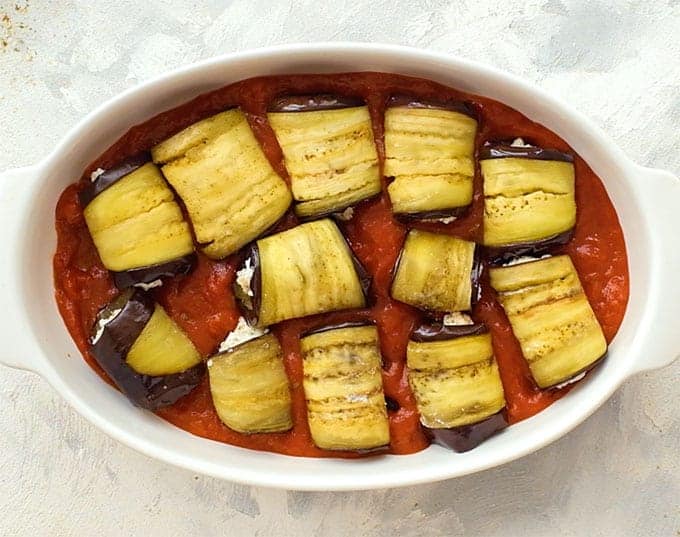 Sliced roasted eggplant rolled around ricotta in a baking dish with marinara sauce