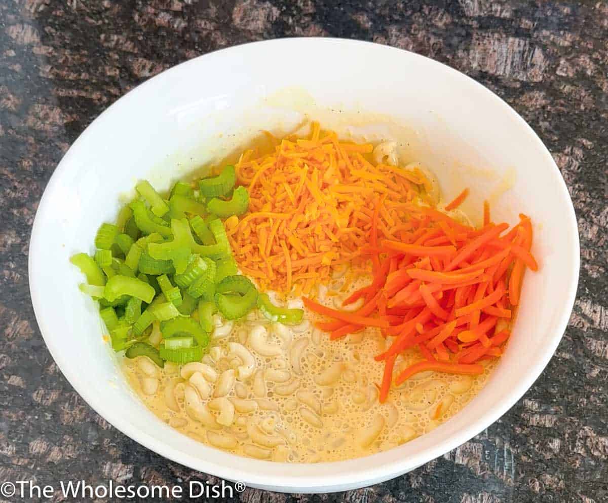 Celery, carrots, and cheese being added to macaroni salad.