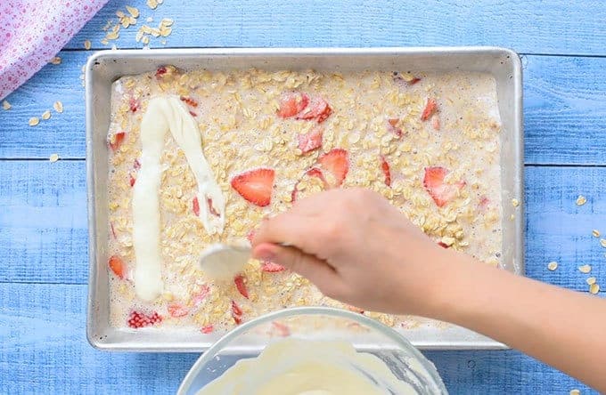 Cheesecake being drizzled over a baking dish full of strawberry baked oatmeal