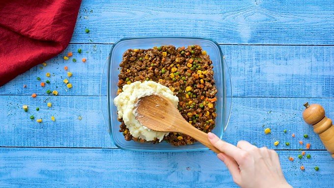 A casserole dish with Cottage Pie beef on the bottom and parmesan mashed potatoes being spread on top