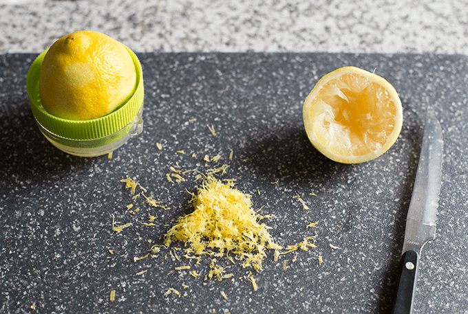 lemon zest, a cut lemon, and a lemon juicer on a dark grey cutting board